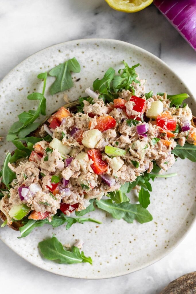 Overhead shot of healthy tuna salad without mayo on top of arugula on a piece of toast on a plate. Next to plate is half a lemon and a red onion.