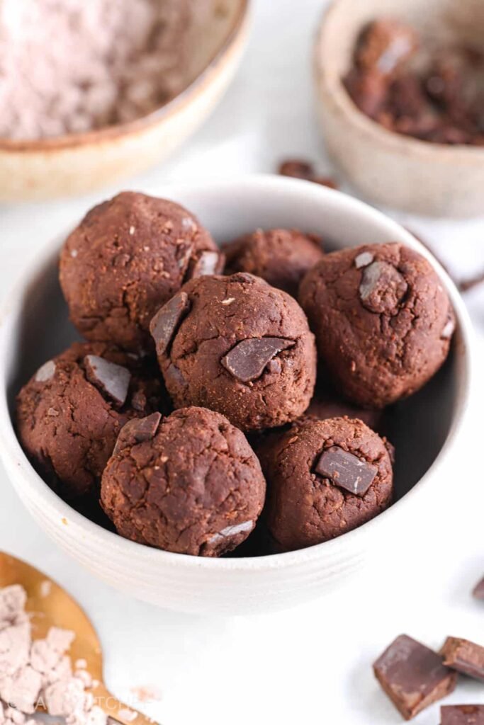 No Bake Protein Brownie Bites {Low Calorie Easy Recipe} Close-up of protein brownie bites with chocolate protein powder and chocolate chunks in the background