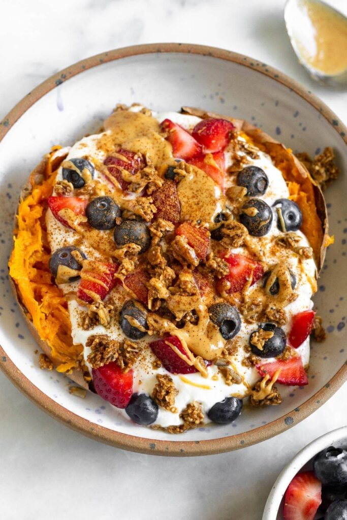 Sweet Potato Breakfast Bowl w/ Greek Yogurt Sweet potato breakfast bowl with greek yogurt, berries, nut butter, granola, and topped with cinnamon. Next to the bowl is a bowl of berries and a spoon with peanut butter on it.