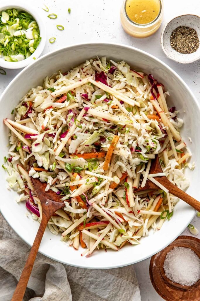 Overhead view of a white bowl filled with colorful Apple Slaw topped with black pepper and green onions.