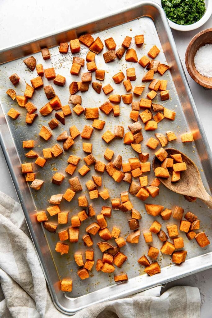 Overhead view of a metal sheet pan filled with freshly Roasted Sweet Potatoes topped with seasonings and crisp on the edges.