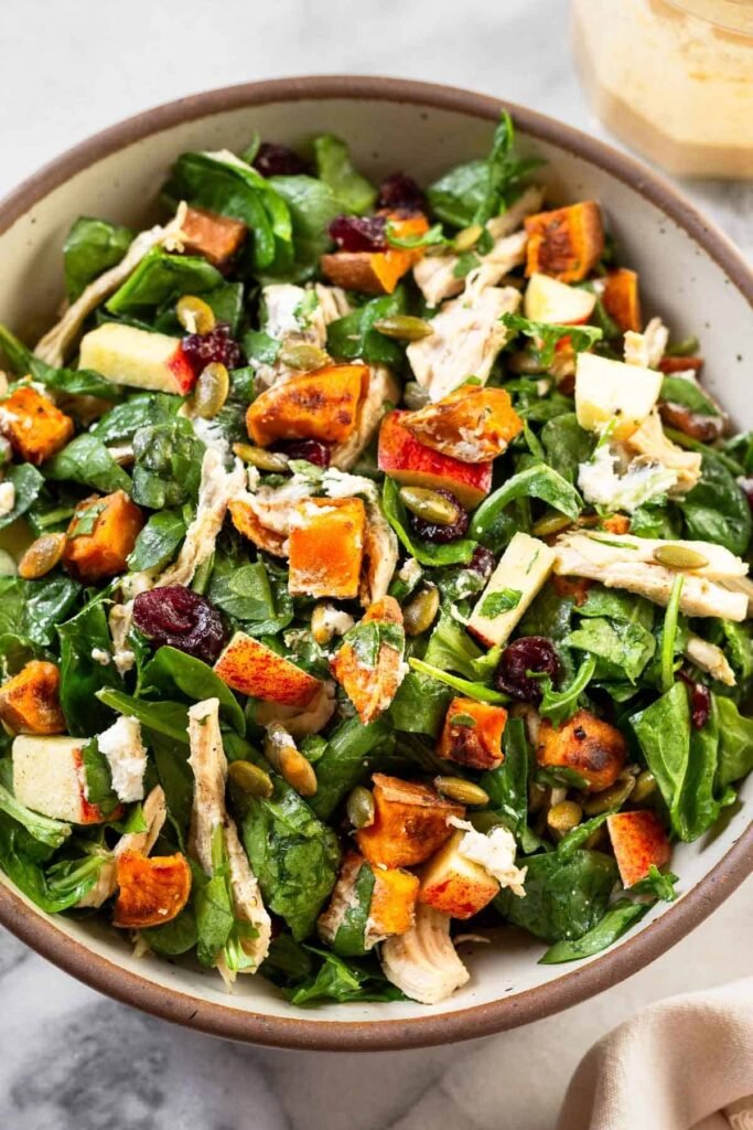 Chopped harvest salad with chicken, mixed greens, roasted sweet potatoes, dried cranberries, pumpkin seeds, and goat cheese all tossed with dressing in a white bowl. Next to the bowl is a jar of more dressing and a white kitchen towel.