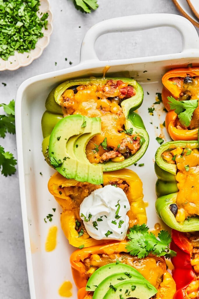 Taco Stuffed Peppers – Eat the Gains Mexican stuffed peppers with ground turkey and melted cheese in a large white casserole dish. They are garnished with sour cream, sliced avocado, and cilantro. Next to the pan is some cilantro leave and a plate of chopped cilantro.