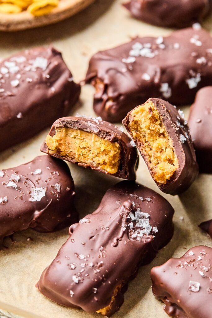 Homemade butterfinger candy bars on a piece of parchment paper. One of the bars is broken in half so you can see the inside of the bar. Off in the corner is a bowl of cornflakes.
