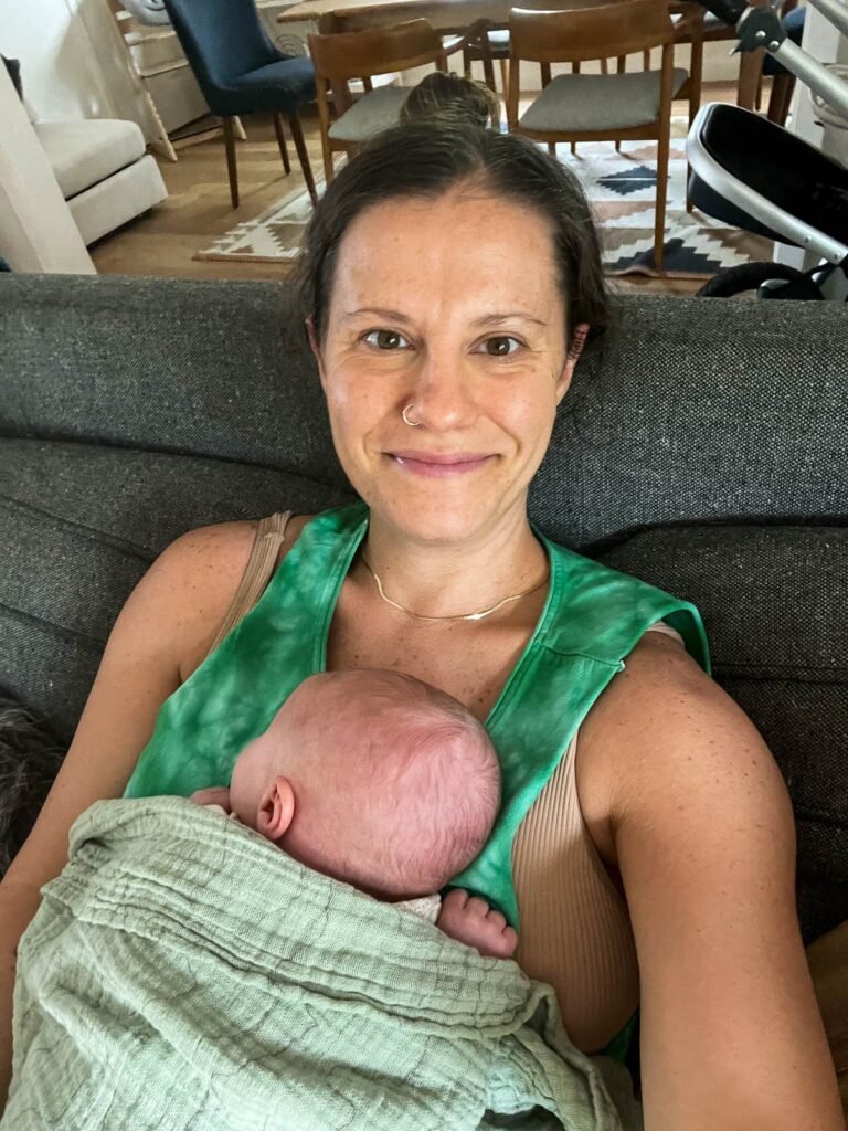 Selfie of a mom sitting on the couch with her baby napping on her. There is a blanket on the baby.