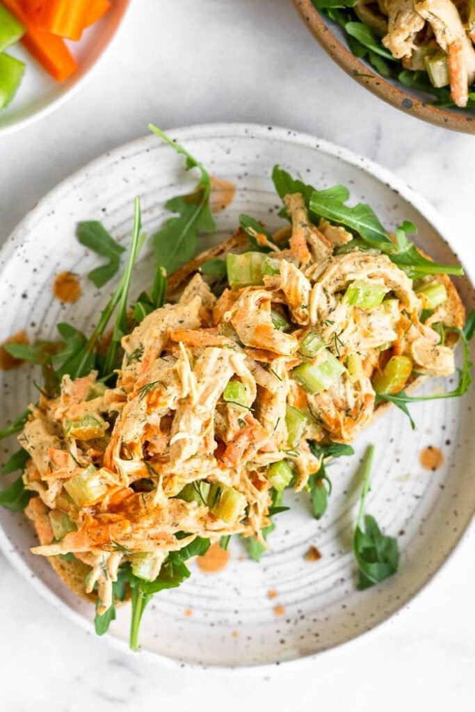 The Best Healthy Chicken Salad Recipes Healthy chicken salad with celery, carrots, and buffalo sauce on top of some arugula on a piece of bread on a plate. It is drizzled with hot sauce. Around the plate is a plate of carrots and celery and a bowl of more chicken salad.