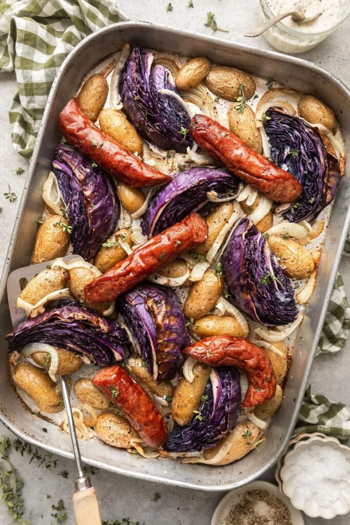 Overhead view of a sheet pan of kielbasa and potatoes with wedges of purple cabbage topped with fresh herbs and black pepper.