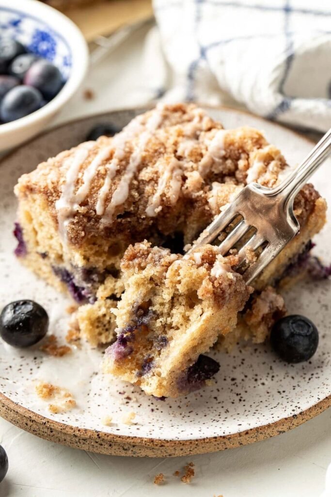 Close up view of a piece of Blueberry Coffee Cake with a fork going into the side drizzled with icing on top.