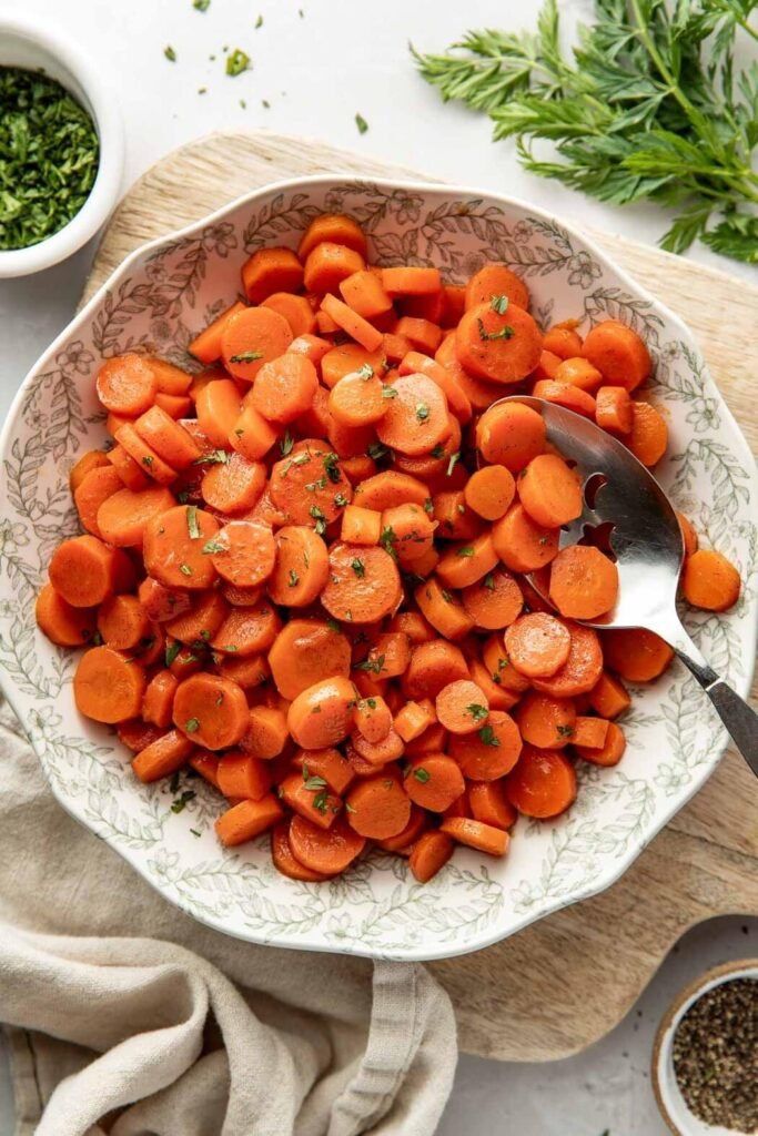Candied Carrots Recipe Overhead view of a decorative bowl filled with sliced cooked carrots topped with fresh herbs and a serving spoon in the side of the bowl.