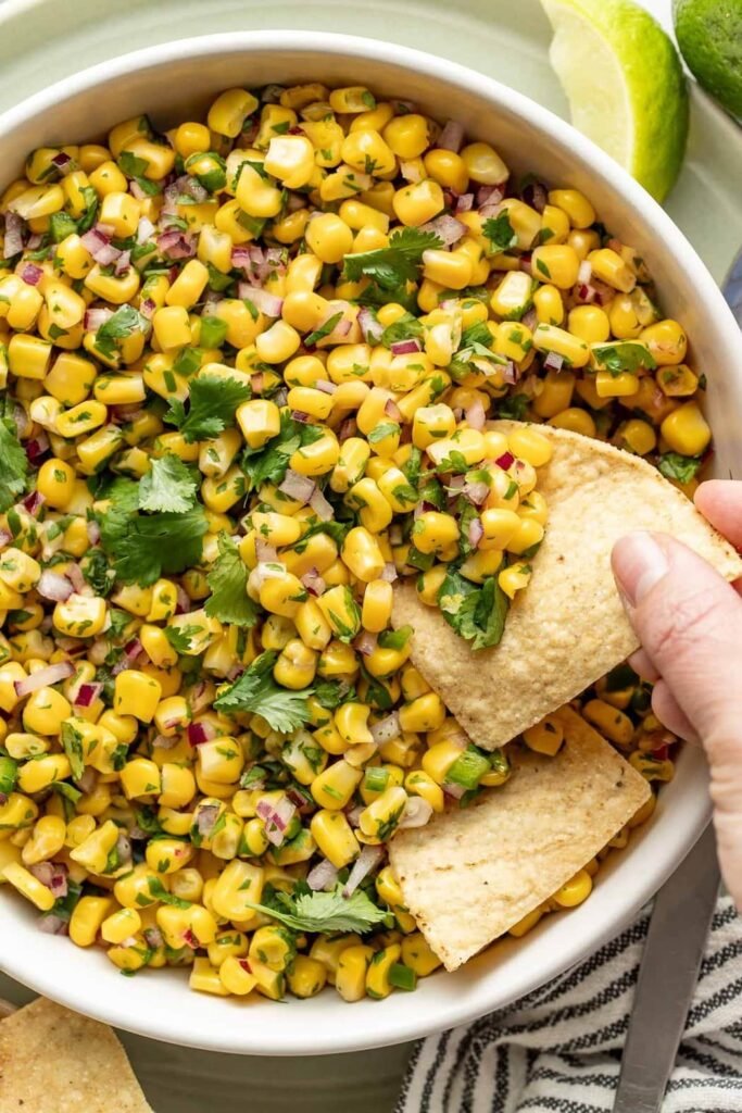 Overhead view of a bowl of Copycat Chipotle Corn Salsa with a hand scooping a tortilla chip into the mixture.