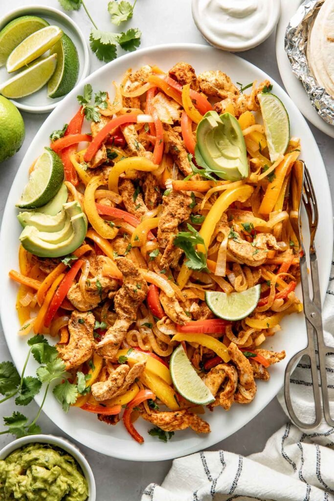 Overhead view of a platter filled with Chicken Fajitas and garnished with avocado, limes, and cilantro.