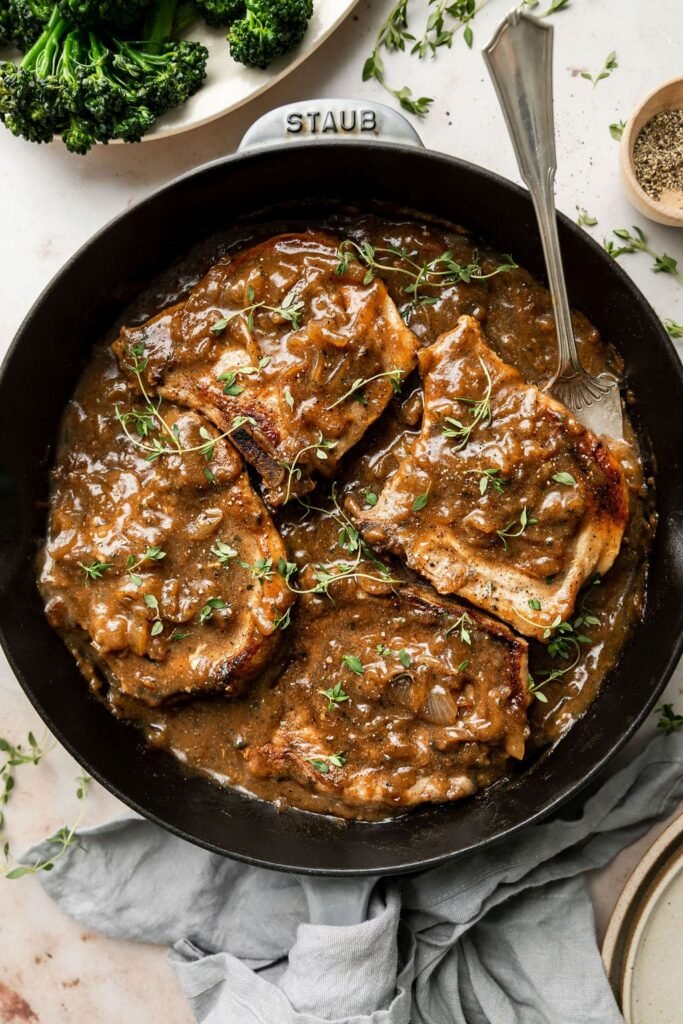 Overhead view of a cast iron pan filled with The Best Smothered Pork Chops topped with fresh herbs and brown gravy.