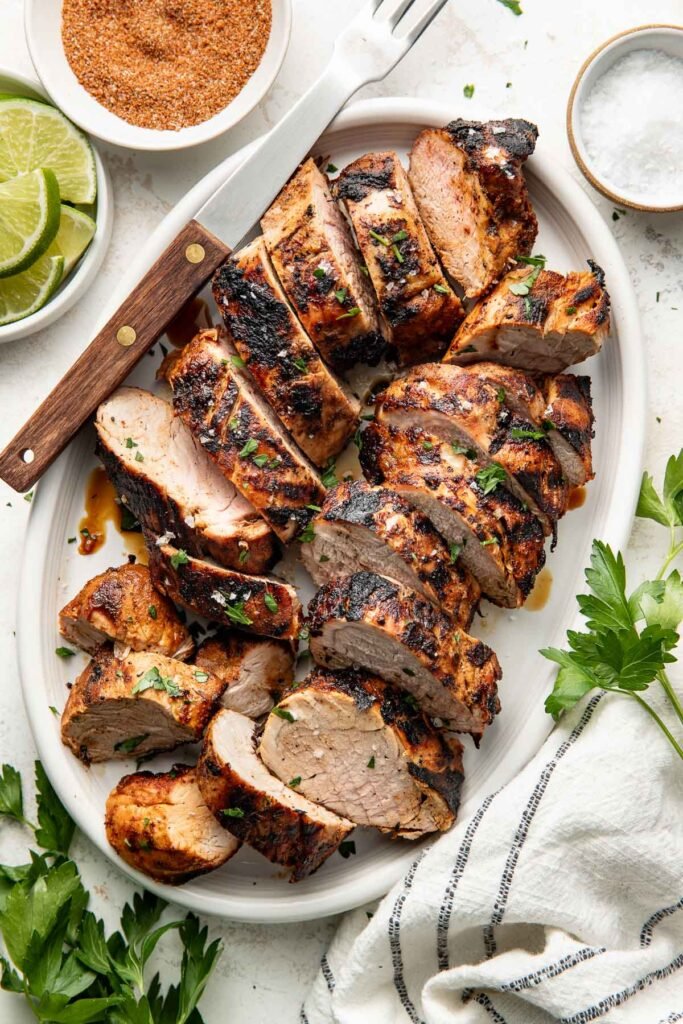 Perfectly grilled and sliced pork tenderloins arranged on a white oval platter surrounded by fresh parsley, limes and red spice mixture in a white bowl.