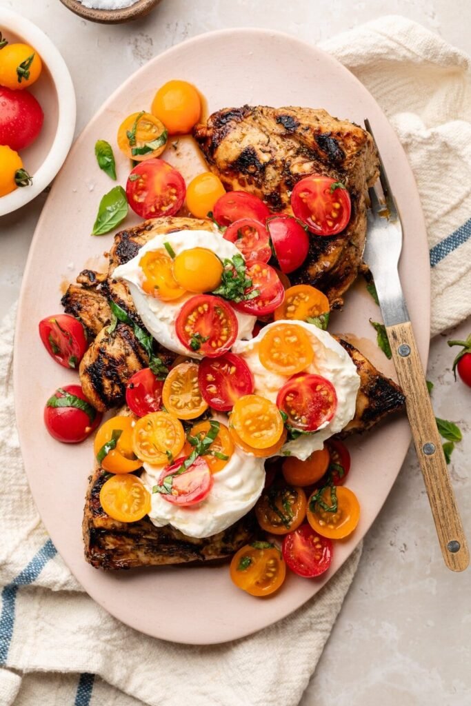 Overhead view of a light pink platter topped with several pieces of grilled chicken breast topped with burrata cheese and a compote of halved red and yellow cherry tomatoes.