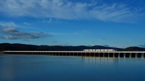 Northern A Northern train on the Furness line crossing the bay at dusk, with the fells in the background. The water and sky appear as a deep blue. The five or six carriage train - mainly white but with blue detailing - is on an extremely long and low viaduct which ploughs a straight line across the wide expanse of water.