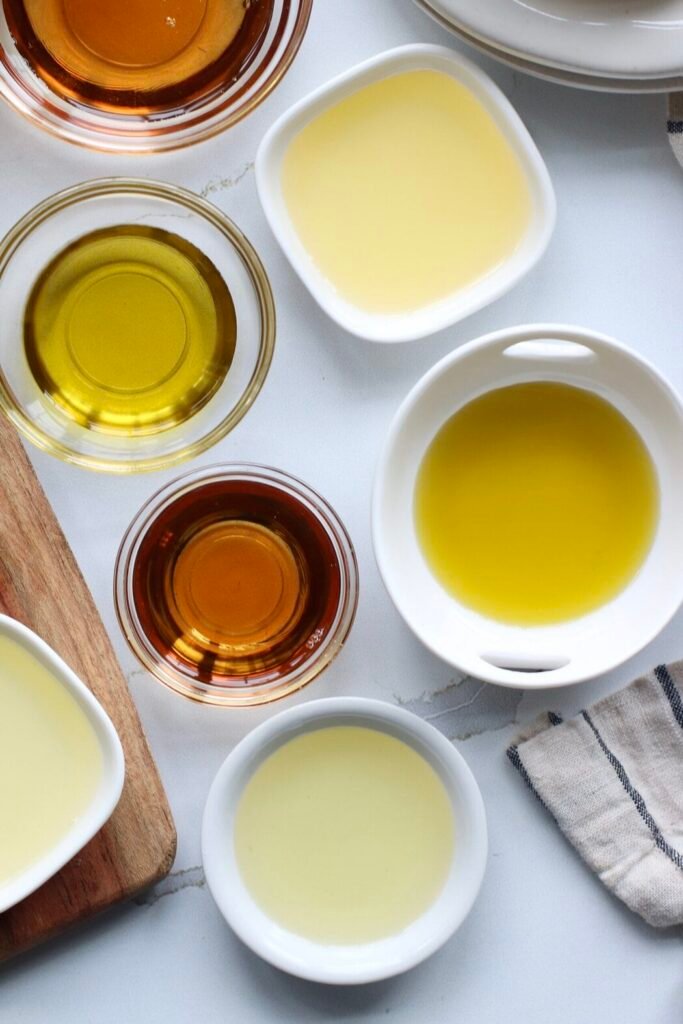 Overhead view of a variety of different colored cooking oils in different sized bowls on a white marble countertop.
