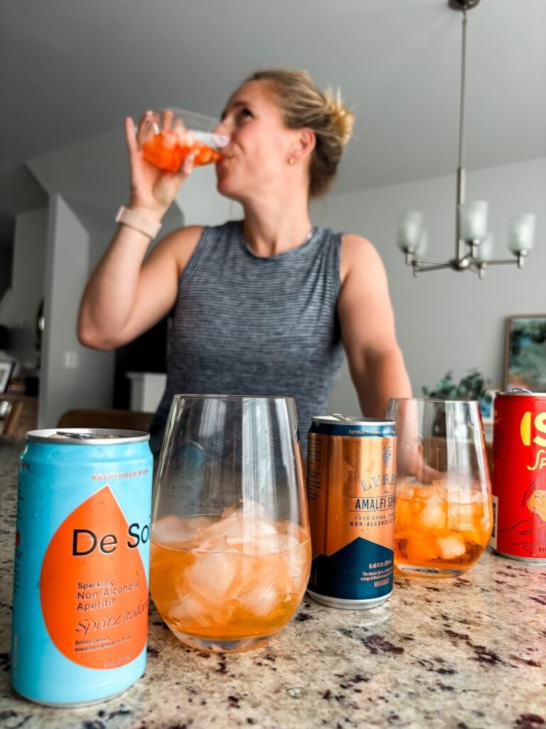 The Best Nonalcoholic Aperol Spritz Options to Try Right Now • Kath Eats Kath drinking a nonalcoholic aperol spritz in the background with cans in the foreground